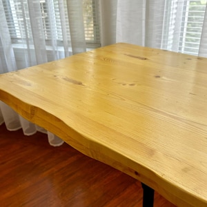 May include: A wooden table top with a live edge design. The table top is light brown and has a smooth, polished finish. The table top is supported by a black metal base with a tripod design.