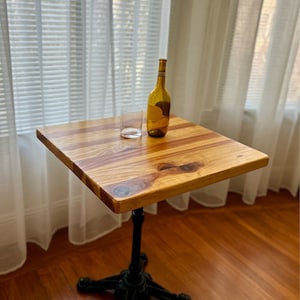 May include: A small wooden table with a black cast iron base. The table top is made of light wood with a natural finish. A glass and a bottle of wine are on the table.