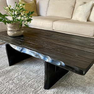 May include: A black wood coffee table with a live edge top and black painted legs. The table is sitting on a white rug in front of a white sofa.