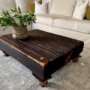 May include: A dark wood coffee table with a distressed finish and metal accents. The table has four wheels and is sitting on a white rug. A vase with white flowers is on the table.