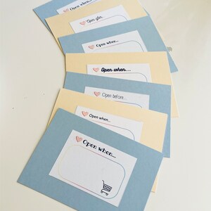 Puede incluir: Un conjunto de siete sobres azules y amarillos con corazones rosas y el texto "Abrir cuando..." en cada sobre. Los sobres están apilados uno encima del otro.