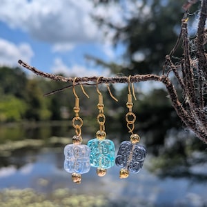 Può includere: Tre paia di orecchini pendenti con ganci e dettagli color oro. Ogni orecchino presenta una perla quadrata e strutturata nelle tonalità di azzurro, turchese e blu scuro. Gli orecchini sono esposti su un piccolo ramo contro uno sfondo sfocato di acqua e cielo.