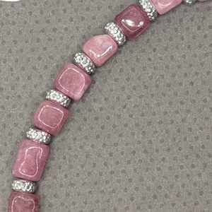 Puede incluir: Primer plano de una pulsera de cuentas rosas. La pulsera presenta cuentas cuadradas de color malva intercaladas con cuentas plateadas con incrustaciones de pedrería. La pulsera está sobre un fondo gris.