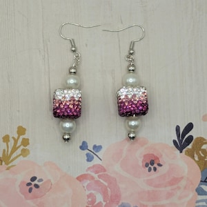 Puede incluir: Un par de pendientes colgantes con ganchos plateados. Cada pendiente presenta una cuenta rectangular con un efecto degradado de cristal rosa y blanco, y está adornado con una cuenta de perla blanca. Los pendientes se muestran sobre un fondo floral.