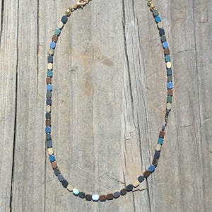 Puede incluir: Un collar de cadena dorada con una serie de pequeñas cuentas cuadradas en tonos de azul, verde, marrón y dorado.