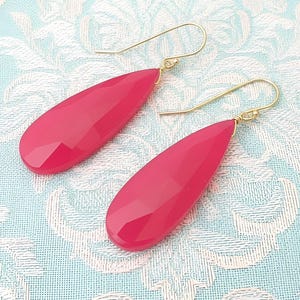 May include: A pair of drop earrings featuring faceted, teardrop-shaped hot pink stones suspended from thin, gold-toned metal ear wires. The stones have a smooth, polished surface with geometric facets that catch the light, creating a vibrant appearance.