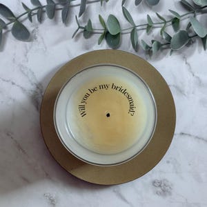May include: A candle in a clear glass container with a gold-colored base. The candle has a light yellow wax and the text "Will you be my bridesmaid?" is printed on the wax in a semi-circle.