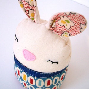 May include: A white fabric bunny with a blue and red patterned fabric bottom. The bunny has pink felt cheeks and embroidered eyes. The bunny's ears are made of a red and white patterned fabric.