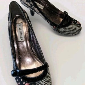 May include: Pair of Steve Madden black and patterned pumps. The shoes feature a mix of patterns, including polka dots, floral, and herringbone, with a glossy black strap and heel. The interior is a metallic gold color.