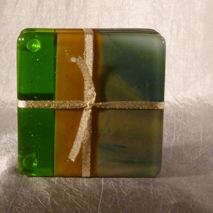 May include: A set of four square glass coasters. Each coaster is divided into three vertical sections of green, gold, and olive green. A gold ribbon is tied around the set. The coasters are a decorative item.