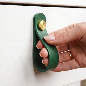 Op de afbeelding: Groene leren ladeknop met een messing knop. De knop is bevestigd aan een witte lade. De afbeelding toont een hand die aan de knop trekt. Een stijlvol woonaccessoire.