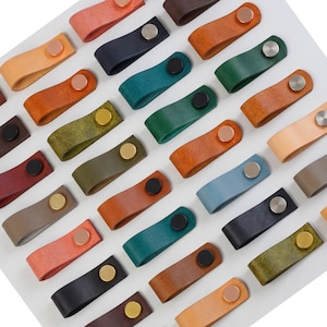 Könnte beinhalten: Eine Auswahl an Leder-Schubladengriffen in verschiedenen Farben, darunter Braun, Orange, Grün und Blau. Jeder Griff hat eine Metallniete in Silber, Gold oder Schwarz. Die Griffe sind auf einer weißen Oberfläche angeordnet.