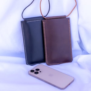 Puede incluir: Dos fundas de teléfono de cuero, una negra y otra marrón, con bordes cosidos y una correa para el cuello. Un smartphone dorado descansa debajo sobre un fondo de tela blanca. Las fundas están diseñadas para guardar un teléfono.