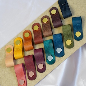 Op de afbeelding: Een verzameling kleurrijke leren lade-trekkers in verschillende tinten, waaronder roze, geel en blauw. Elke trekker heeft een metalen klinknagel en een goudkleurig cirkelvormig accent. De trekkers zijn gerangschikt op een lichtbruin oppervlak.