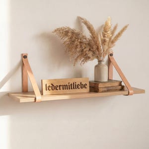 Op de afbeelding: Een licht houten zwevend rek met leren banden, met een kleine vaas met gedroogde planten, boeken en een houten bord met de tekst "ledermitliebe". Het rek is aan een neutraal gekleurde muur bevestigd.