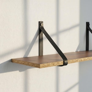 Può includere: Una mensola sospesa con superficie in legno marrone scuro e supporti in cinturino di pelle nera. La mensola è montata su una parete bianca e il design è minimalista e moderno. La mensola è lunga circa 76 cm.