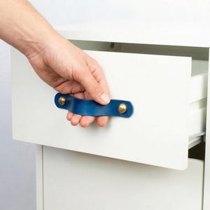 Può includere: Una maniglia per cassetto in pelle blu con rivetti in ottone è attaccata a un cassetto bianco. Il cassetto è parzialmente aperto, rivelando l'interno. La maniglia è curva e ha un design semplice e moderno.