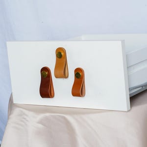 Peut inclure: Trois poignées de tiroir en cuir de différentes nuances de marron sont fixées sur une surface blanche. Chaque poignée est constituée d'une boucle de cuir fixée par un rivet en laiton. Les poignées sont disposées horizontalement.