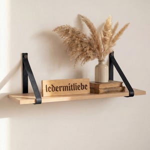 Op de afbeelding: Een zwevend houten plankje met zwarte leren banden, met een beige vaas met gedroogd pampasgras, een stapel boeken en een houten bord met de tekst "ledermitliebe". Het plankje is aan een lichtbeige muur bevestigd.