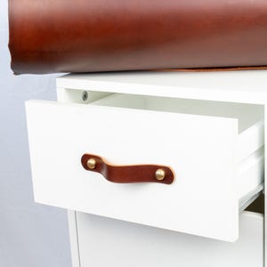 Op de afbeelding: Witte kast met een open lade met een bruine leren handgreep met messing accenten. Een grote rol bruin leer ligt bovenop de kast. De kast heeft een modern design.