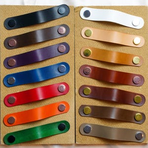 Puede incluir: Una colección de tiradores de cajón de cuero en varios colores, incluyendo negro, blanco, marrón, morado, azul, rojo, naranja y verde. Cada tirador está sujeto con remaches metálicos. Los tiradores están dispuestos sobre una superficie beige.
