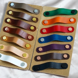 Könnte beinhalten: Eine Sammlung farbenfroher Leder-Schubladengriffe in verschiedenen Farbtönen, darunter Braun, Grün, Orange, Rot, Blau, Lila und Weiß. Jeder Griff ist mit Metallnieten befestigt. Die Griffe sind auf zwei rechteckigen Karton-Displays angeordnet.