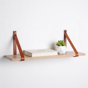 Peut inclure: Une étagère en bois clair avec des lanières en cuir marron, supportant un livre fermé et une petite plante succulente en pot. L'étagère est fixée à un mur blanc, créant une esthétique minimaliste.
