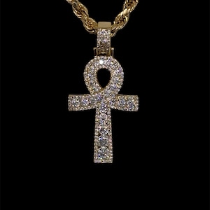 May include: A gold-colored pendant necklace featuring an Ankh cross design, encrusted with numerous small, sparkling gemstones. The pendant hangs from a thick, twisted rope chain, also gold-toned, against a black background.