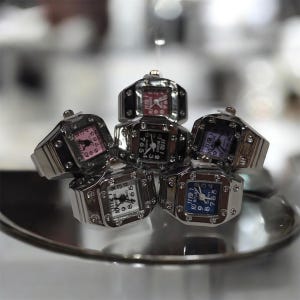 May include: A collection of silver-toned ring watches with square faces and various colored dials, including pink, black, white, and blue. The rings are arranged on a reflective surface, showcasing the timepieces' details.