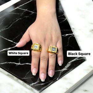 Puede incluir: Dos anillos cuadrados dorados con esferas de reloj, uno con un cuadrado blanco y otro con un cuadrado negro. Los anillos est&aacute;n en una mano, con el texto "White Square" y "Black Square" visibles.