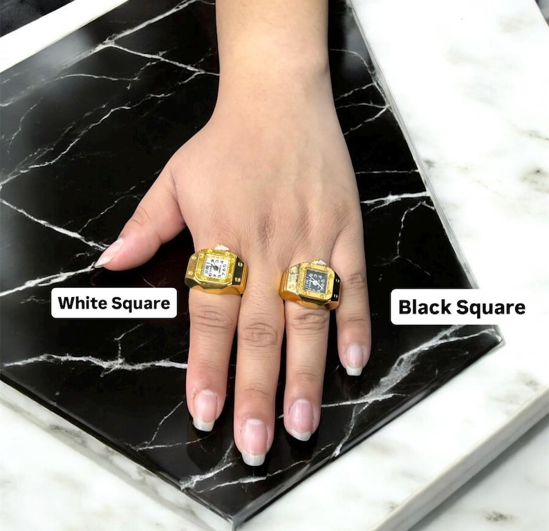Puede incluir: Dos anillos cuadrados dorados con esferas de reloj, uno con un cuadrado blanco y otro con un cuadrado negro. Los anillos est&aacute;n en una mano, con el texto "White Square" y "Black Square" visibles.