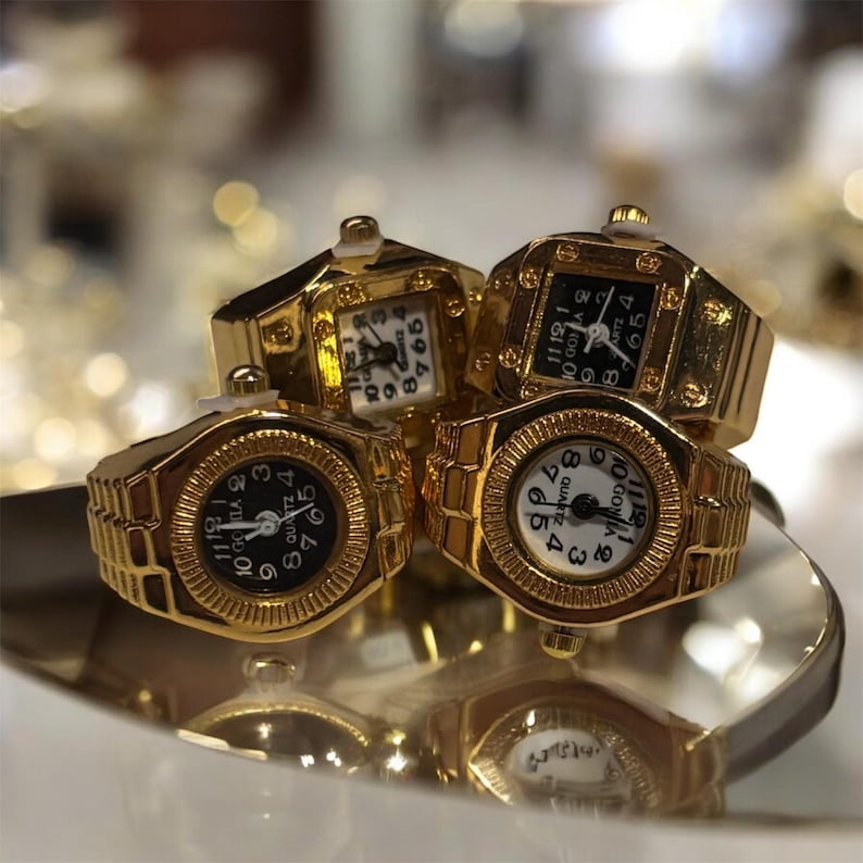 Puede incluir: Cuatro anillos-reloj dorados con esferas en blanco y negro. Los anillos tienen un dise&ntilde;o de esfera rectangular o circular con un anillo tipo banda. Los relojes se exhiben sobre una superficie reflectante.