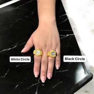 Puede incluir: Dos anillos dorados con forma de reloj, uno con esfera blanca y otro con esfera negra, se muestran en una mano. Los anillos est&aacute;n sobre una superficie de m&aacute;rmol blanco y negro. El texto "White Circle" y "Black Circle" son visibles.
