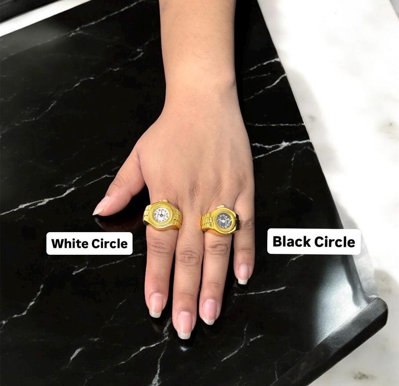 Puede incluir: Dos anillos dorados con forma de reloj, uno con esfera blanca y otro con esfera negra, se muestran en una mano. Los anillos est&aacute;n sobre una superficie de m&aacute;rmol blanco y negro. El texto "White Circle" y "Black Circle" son visibles.