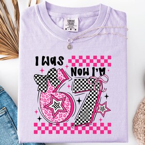 May include: A light purple Comfort Colors t-shirt with the text "I WAS" above the number "57" and "NOW I'M" above the number "57". The numbers are pink with a leopard print and a checkered pattern. A silver necklace is on the shirt.
