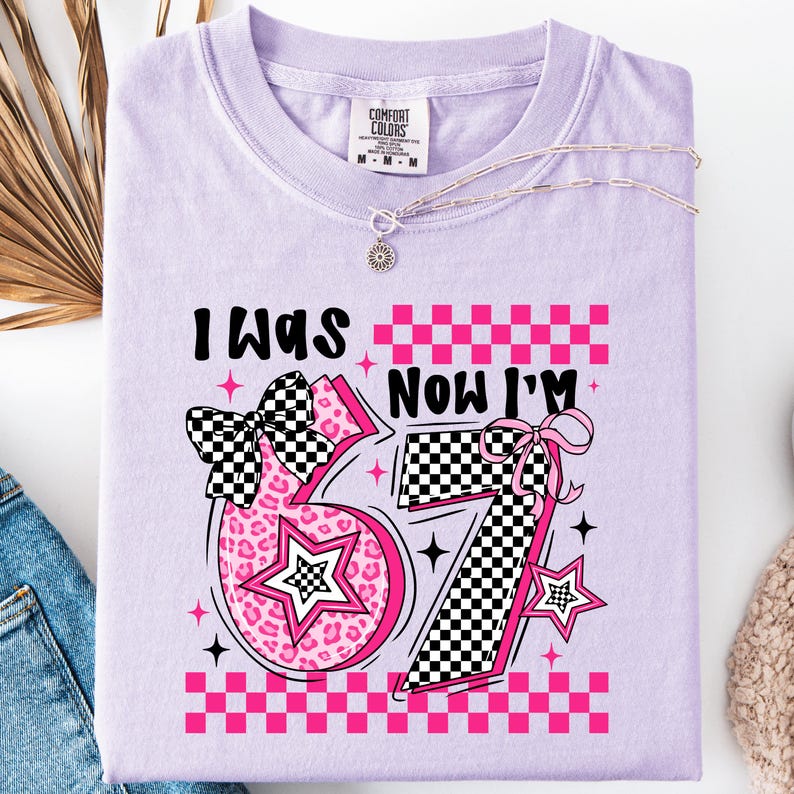May include: A light purple Comfort Colors t-shirt with the text "I WAS" above the number "57" and "NOW I'M" above the number "57". The numbers are pink with a leopard print and a checkered pattern. A silver necklace is on the shirt.