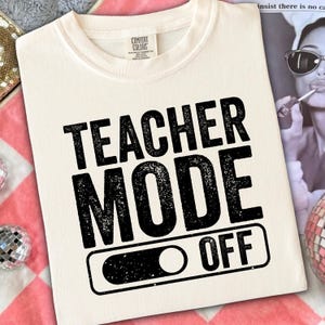May include: Cream-colored t-shirt with the words "TEACHER MODE OFF" in bold, black, distressed font. The design includes an on/off switch graphic. The shirt is made by Comfort Colors.