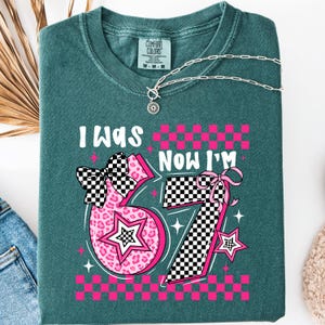 May include: Teal Comfort Colors t-shirt with the text "I WAS" above the number "67" and "NOW I'M" above the number "67". The numbers are pink with a star and bow design. A silver necklace is draped across the shirt.