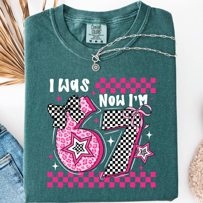 May include: Teal Comfort Colors t-shirt with the text "I WAS" above the number "67" and "NOW I'M" above the number "67". The numbers are pink with a star and bow design. A silver necklace is draped across the shirt.