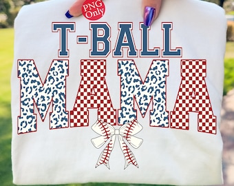 May include: White t-shirt with the text "T-BALL MAMA" in red and blue lettering. The word "MAMA" has a leopard print design. A baseball bow graphic is at the bottom. The shirt is held up by a hand.