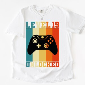 May include: White t-shirt with a graphic featuring a black video game controller and the text "LEVEL 19 UNLOCKED." The design includes vertical stripes in teal, yellow, orange, and red. A silver necklace is visible.