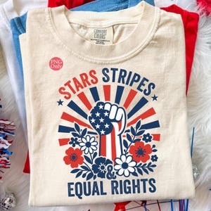 以下が含まれることがあります： クリーム色のTシャツ。星条旗のデザインの握りこぶし、赤、白、青の光線、花、そして「STARS STRIPES & EQUAL RIGHTS」の文字が描かれた愛国的なデザインです。