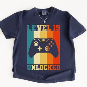 May include: Navy blue t-shirt with a retro design featuring a video game controller and the text "LEVEL 19 UNLOCKED" in a colorful, vertical striped pattern. The shirt has short sleeves and a crew neck.