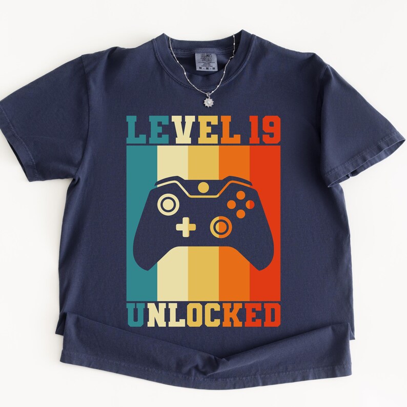 May include: Navy blue t-shirt with a retro design featuring a video game controller and the text "LEVEL 19 UNLOCKED" in a colorful, vertical striped pattern. The shirt has short sleeves and a crew neck.