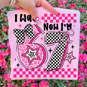 May include: A pink sweatshirt featuring the text "I Was 6 Now I'm 7". The design includes a leopard print number 6, a star, and a checkered bow. The number 7 has a checkered pattern and a pink bow, with a pink checkered border.