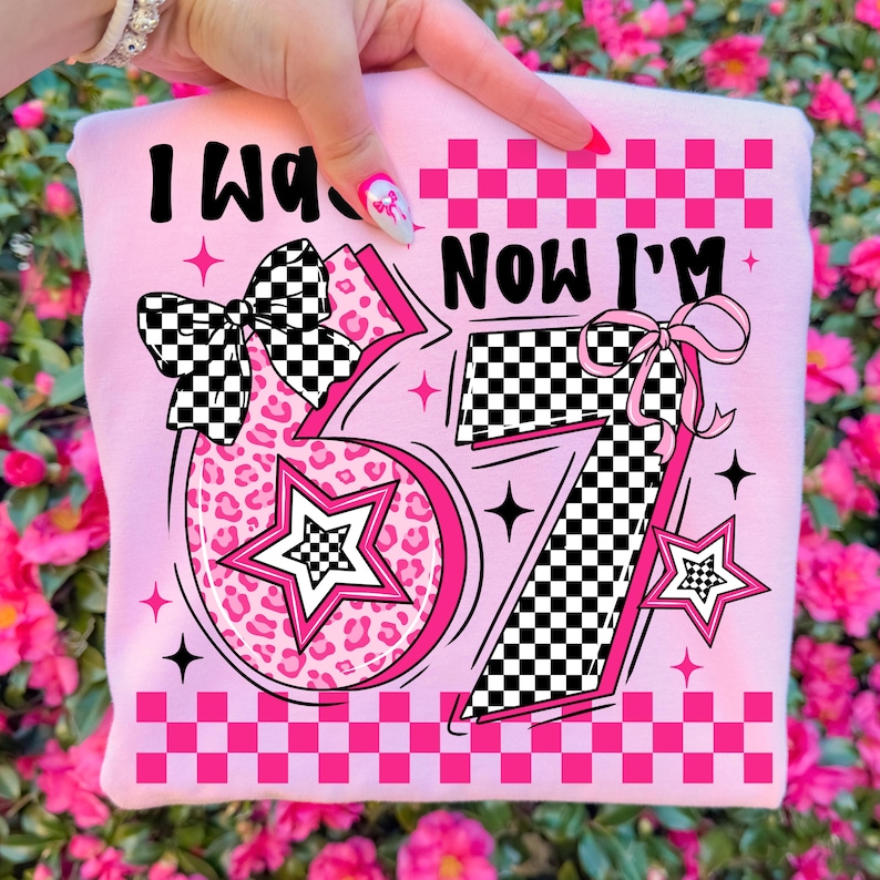 May include: A pink sweatshirt featuring the text "I Was 6 Now I'm 7". The design includes a leopard print number 6, a star, and a checkered bow. The number 7 has a checkered pattern and a pink bow, with a pink checkered border.