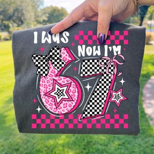May include: Dark grey t-shirt featuring the text "I was... Now I'm 57". The number 5 is pink with a leopard print, and the number 7 is black and white checkered. The design includes stars and bows.