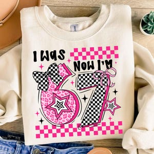 May include: Cream-coloured sweatshirt with the text "I WAS NOW I'M 57". The number 57 is in pink with a leopard print design and a checkered bow. The design includes stars and a checkered pattern.