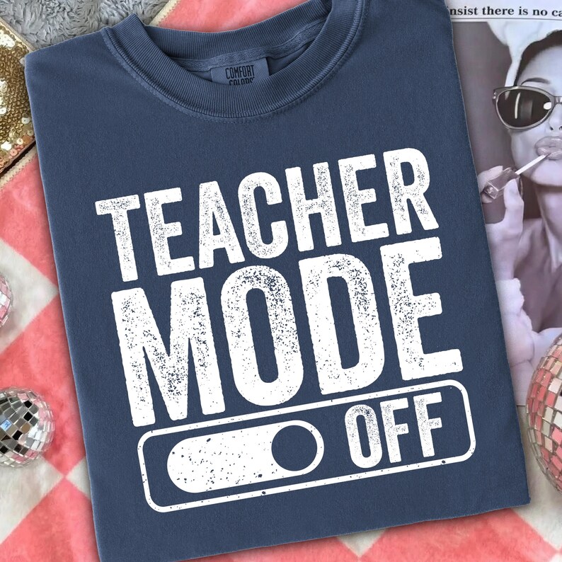 May include: A navy blue t-shirt with the words "TEACHER MODE OFF" in distressed white lettering. The shirt features a switch graphic below the text. The shirt is made of a soft material.