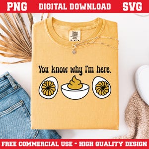 Può includere: T-shirt giallo senape con la scritta "You know why I'm here" e una grafica di uova alla diavola. La maglietta ha girocollo e maniche corte. Una collana d'oro è drappeggiata sulla maglietta. La maglietta è piegata su una superficie bianca.
